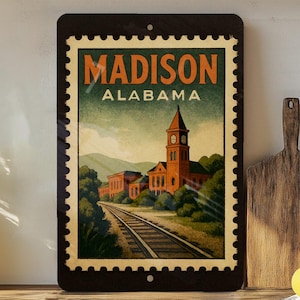 A framed vintage-style poster of Madison, Alabama, featuring a train track and a clock tower, is displayed on a shelf next to a wooden cutting board and a potted plant.