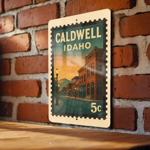 A metal sign with a vintage-style illustration of a street scene in Caldwell, Idaho, mounted on a brick wall.
