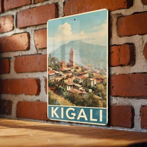 May include: A rectangular metal sign featuring a vintage-style illustration of Kigali, Rwanda. The artwork depicts a cityscape with a prominent church tower, set against a mountain backdrop and a blue sky. The word "KIGALI" is printed at the bottom.