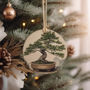 May include: A round, cream-colored Christmas ornament with a detailed illustration of a bonsai tree in a brown pot. The tree has green leaves and a thick, gnarled trunk. The background is a light brown with a faint mountain scene.