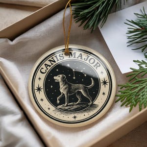May include: A round, cream-colored ornament with a black and white illustration of a dog and the Canis Major constellation. The ornament has the words "CANIS MAJOR" around the edge and is suspended by a gold string. The ornament is in a gift box.