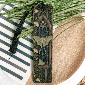 May include: A rectangular bookmark featuring a detailed illustration of a pale green luna moth centered against a dark blue gothic arched window. The design includes climbing vines with small white flowers and a black tassel attached at the top.