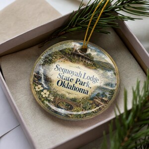 May include: A round Christmas ornament featuring a scenic illustration of Sequoyah Lodge State Park, Oklahoma. The ornament has a gold string for hanging and is presented in a gift box with a sprig of evergreen.