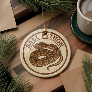 May include: A round, cream-colored ornament with a brown and tan ball python illustration. The words "BALL PYTHON" are printed in a circular design around the snake. The ornament has a gold string for hanging.