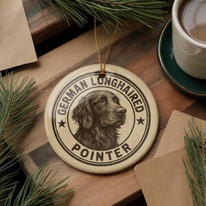 May include: Cream-colored round ornament with a German Longhaired Pointer illustration. The ornament has the text "GERMAN LONGHAIRED POINTER" in a circular design. A gold string is attached for hanging. The ornament is on a wooden surface.