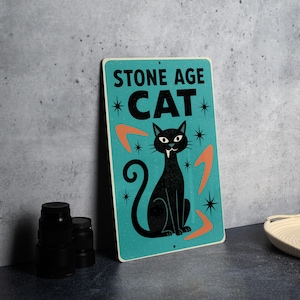 May include: A retro-style metal sign with the words "STONE AGE CAT" in black. The sign features a black cat illustration, orange boomerangs, and starbursts on a teal background. The sign has a vintage aesthetic.