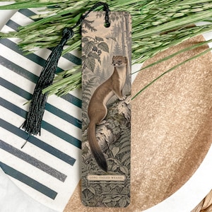 May include: A rectangular bookmark featuring a vintage-style illustration of a long-tailed weasel standing on a log amidst forest foliage. The bookmark has a black tassel attached to the top and the text LONG-TAILED WEASEL printed at the bottom.