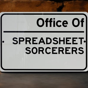 May include: White sign with black text that reads "Office Of . SPREADSHEET. SORCERERS".