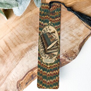 May include: A rectangular bookmark featuring a multi-colored marbled paper pattern in shades of green, red, and gold. The center displays an oval illustration of a book with a stylus and the text MARBLED PAPER BOOKBINDING. A black tassel is attached.