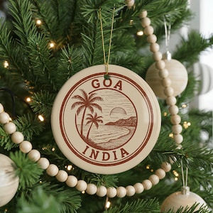 May include: A round, cream-colored Christmas ornament with a brown illustration of a beach scene with palm trees and the text "GOA INDIA". The ornament hangs from a gold string and is surrounded by a Christmas tree.