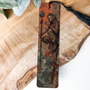 May include: A bookmark featuring a detailed illustration of a person holding a staff with a circular orb on top. The person is standing on a rocky cliff with a red and orange sky in the background. The bookmark is made of metal and has a black tassel.