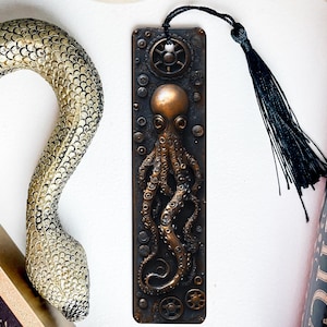 May include: A bronze bookmark with a steampunk octopus design. The bookmark has a black tassel and is made of metal.