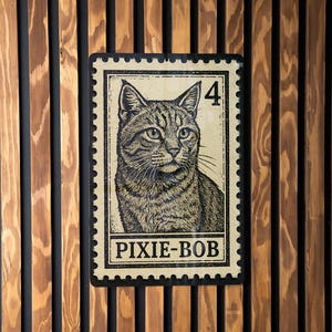 May include: A rectangular sign featuring a detailed illustration of a cat, resembling a postage stamp. The sign has a cream-colored background with the text "PIXIE-BOB" and the number "4". The sign is mounted on a wooden wall with vertical slats.