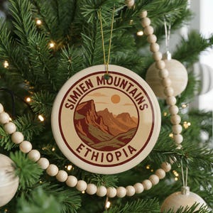 May include: A round ceramic Christmas ornament featuring a landscape design of the Simien Mountains in Ethiopia. The ornament has a cream-colored background with a brown and orange mountain scene and the words "Simien Mountains Ethiopia" in a circular design.