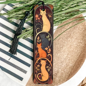 May include: A black bookmark with a gold trim features three cats in a stylized design. The cats are orange, black, and white and are surrounded by floral motifs. The bookmark has a black tassel.