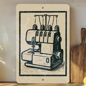 May include: A rectangular sign featuring a black and white illustration of a serger sewing machine. The serger is depicted with multiple thread spools and a detailed mechanical design. The sign has a cream-colored background and a black border.