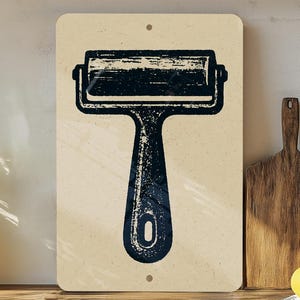 May include: A rectangular sign featuring a dark blue ink roller design. The roller is centered on a beige background with a distressed, vintage aesthetic. The sign has two small holes for hanging.