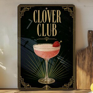 May include: A decorative sign featuring a pink cocktail with a raspberry garnish, labeled "CLOVER CLUB" in gold lettering. The sign has a black background with art deco style gold borders and accents. The image is set against a neutral background with a wooden cutting board and lemons.