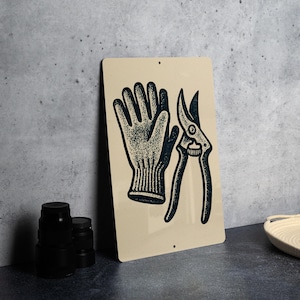 May include: Tan metal sign featuring a black illustration of a gardening glove and pruning shears. The glove has a ribbed cuff and detailed stitching. The shears are open, showing the blades and handle. The sign has two small holes for hanging.