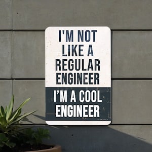 May include: A white sign with black text that reads "I'm not like a regular engineer. I'm a cool engineer." The sign is mounted on a gray wall.