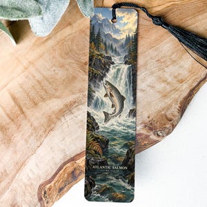 May include: A rectangular bookmark featuring a detailed illustration of an Atlantic salmon leaping up a waterfall surrounded by pine trees and mountains. The bookmark has a black tassel attached to the top and the text Atlantic Salmon printed at the bottom.