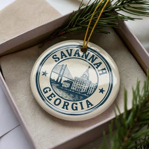 May include: A round, cream-colored ornament with a blue design featuring the text "SAVANNAH GEORGIA" in a circular border. The design includes a bridge, buildings, and stars. The ornament is in a gift box with a gold string.
