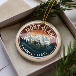 May include: Cream-colored round ornament with a mountain scene and the words "MONT BLANC FRANCE ITALY" in a circular design. The ornament is in a gift box with a gold string for hanging and a sprig of pine.