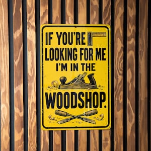 May include: A rectangular yellow metal sign with black text that reads IF YOU'RE LOOKING FOR ME I'M IN THE WOODSHOP. The sign features a graphic illustration of a hand plane and two crossed chisels, with small wood shavings scattered around.