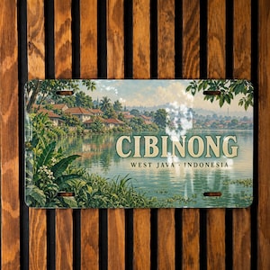 May include: A rectangular metal sign featuring a scenic landscape illustration of a lake with houses and lush greenery. The text CIBINONG WEST JAVA INDONESIA is printed in the center. The sign has four mounting holes, one in each corner.