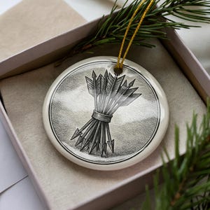 May include: A round, cream-colored Christmas ornament with a black and white illustration of a bundle of arrows. The ornament is inside a pink gift box with a gold string for hanging. Green pine branches are in the background.