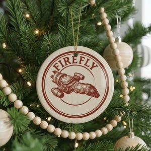 May include: A round, off-white Christmas ornament with a red border and the word "FIREFLY" in a circular design. A red illustration of a spaceship is in the center. The ornament hangs from a gold string on a Christmas tree.