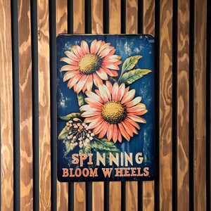 May include: A vintage-style metal sign with a blue background and two pink and orange sunflowers. The sign reads "Spinning Bloom Wheels".