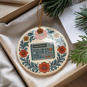 May include: Round ceramic ornament featuring a calculator design. The ornament is cream-colored with a floral border in orange, pink, and teal. A gold string is attached for hanging. The ornament is presented in a gift box.