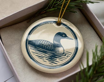 May include: A round, cream-colored ornament featuring a dark blue illustration of a loon swimming in water. The loon is inside a dark blue circular border. The ornament is in a pink box with a gold string and pine needles.