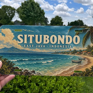 May include: A rectangular metal license plate featuring a scenic illustration of a tropical beach in Situbondo, East Java, Indonesia. The design includes blue ocean waves, a sandy shore, palm trees, and mountains under a cloudy sky.
