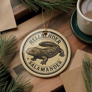 May include: Round ceramic ornament with a tan background and a black and white illustration of a hellbender salamander. The text "HELLBENDER SALAMANDER" is printed around the salamander in a circular design. A gold string is attached for hanging.