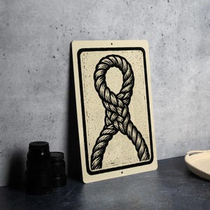 May include: A rectangular metal sign with a black rope design on a cream background. The rope is intricately woven, creating a knot-like pattern. The sign has a black border and two small holes for hanging. The sign is leaning against a gray wall.