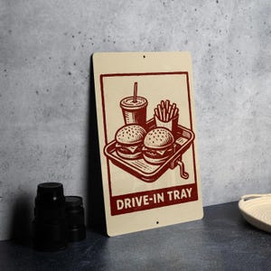 May include: A rectangular cream-colored metal sign with a maroon illustration of a drive-in tray. The tray holds two burgers, a drink, and fries. The words "DRIVE-IN TRAY" are printed in maroon at the bottom of the sign.