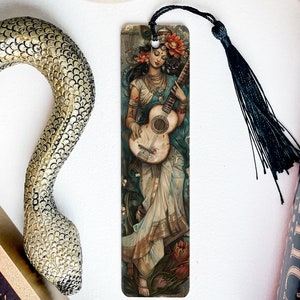 May include: A bookmark featuring a woman playing a guitar. She is wearing a blue and white dress with floral accents. The bookmark is made of sturdy cardstock and has a black tassel.