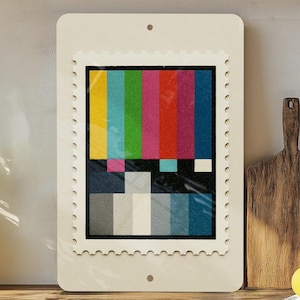 May include: A rectangular, off-white wooden sign with a postage stamp design featuring a colorful test pattern. The sign is displayed on a wooden surface with a plant, cutting boards, and lemons in the background.