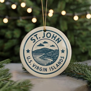 May include: A round, off-white ceramic ornament with a blue illustration of a coastal landscape and the text "ST. JOHN U.S. VIRGIN ISLANDS". The ornament is hanging by a gold string, with a blurred background of Christmas lights and greenery.