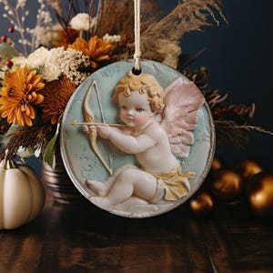 May include: A round ceramic ornament featuring a cherub with pink wings holding a bow and arrow. The cherub is painted in a realistic style with delicate details. The ornament has a light blue background with a white border.