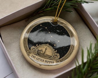 May include: A round, cream-colored ornament featuring a black and white illustration of the Little Dipper constellation. The ornament has a gold string for hanging and is presented in a pink gift box with a sprig of evergreen.