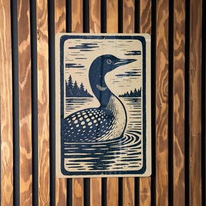 May include: A rectangular print featuring a stylized illustration of a loon swimming in water. The loon is depicted with a dark blue head and back, white spotted chest, and a long, pointed beak. The background shows a lake and trees.