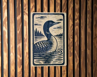 May include: A rectangular print featuring a stylized illustration of a loon swimming in water. The loon is depicted with a dark blue head and back, white spotted chest, and a long, pointed beak. The background shows a lake and trees.