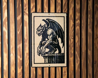 May include: A rectangular metal sign featuring a black and white illustration of a gargoyle with large wings, perched on a stone structure. The sign is framed in black and mounted on a wooden slatted wall.