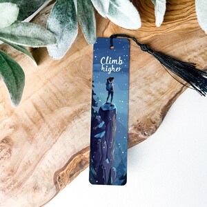 May include: A blue and white bookmark with a woman standing on a mountain top. The text on the bookmark reads "Climb higher".