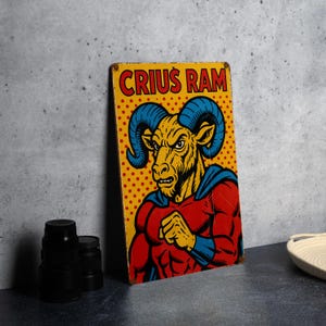 May include: A rectangular metal sign with the text "CRIUS RAM" in red letters. The sign features a muscular ram character with blue horns and a red and blue superhero outfit, set against a yellow background with red dots.