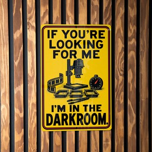 May include: A rectangular yellow metal sign with black text that reads IF YOU'RE LOOKING FOR ME I'M IN THE DARKROOM. The graphic features a black illustration of a film enlarger, a strip of film, a timer, three developing trays, and tongs.