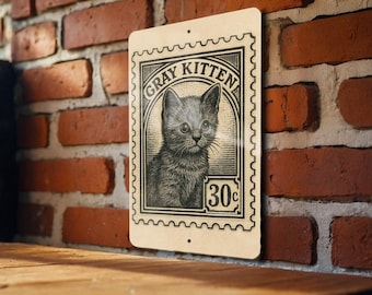 May include: A vintage-style metal sign featuring a gray kitten illustration within a postage stamp design. The sign reads "GRAY KITTEN" and "30¢". The sign is mounted on a red brick wall.